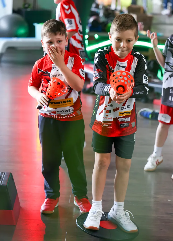 Zwei Jungen im Dart-Team-Outfit, einer bedeckt den Mund mit der Hand, während der andere eine orangefarbene Scheibe hält. Im Hintergrund sind weitere Kinder und Dartscheiben zu sehen.