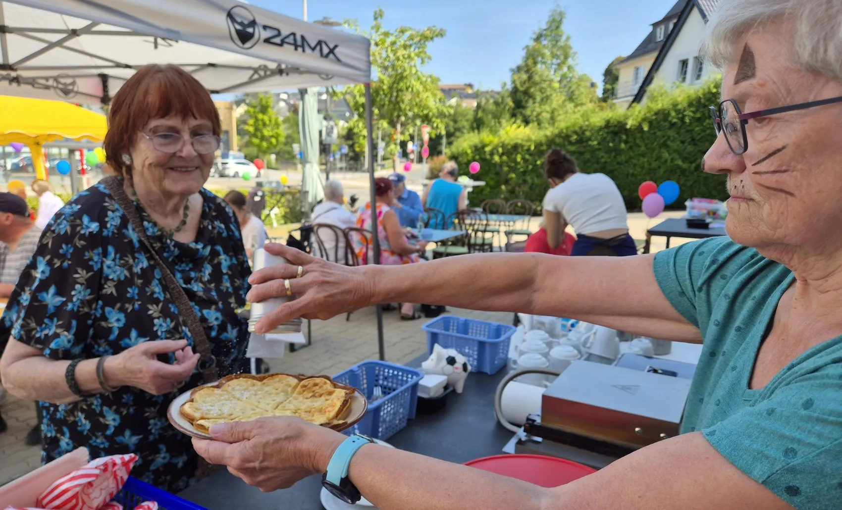 Eine Frau mit aufgemalten Schnurrhaaren reicht einer lächelnden Frau einen Teller mit Gebäck bei einem Sommerfest im Freien.