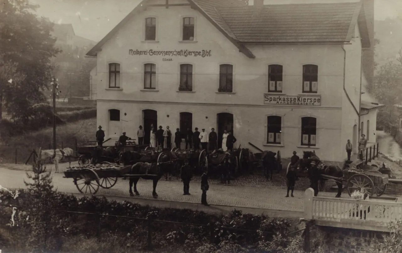Historisches Schwarzweißfoto des ersten Gebäudes der Volksbank Kierspe, das auch als Molkereigenossenschaft und Sparkasse diente. Vor dem Gebäude stehen Menschen und Pferdegespanne.
