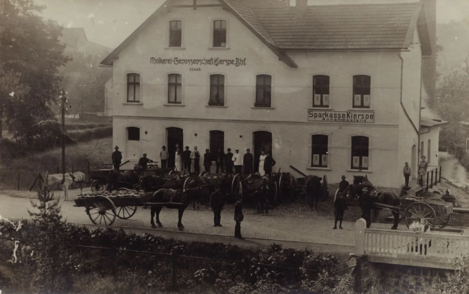 Historisches Schwarzweißfoto des ersten Gebäudes der Volksbank Kierspe, das auch als Molkereigenossenschaft und Sparkasse diente. Vor dem Gebäude stehen Menschen und Pferdegespanne.