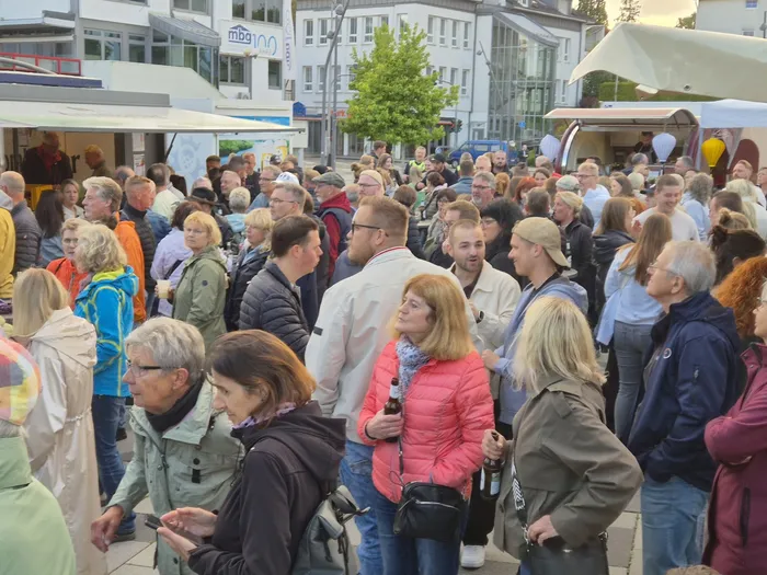 Eine große Menschenmenge steht und geht auf einem Stadtplatz, während im Hintergrund Gebäude und Bäume zu sehen sind. Einige Personen halten Getränke in der Hand.