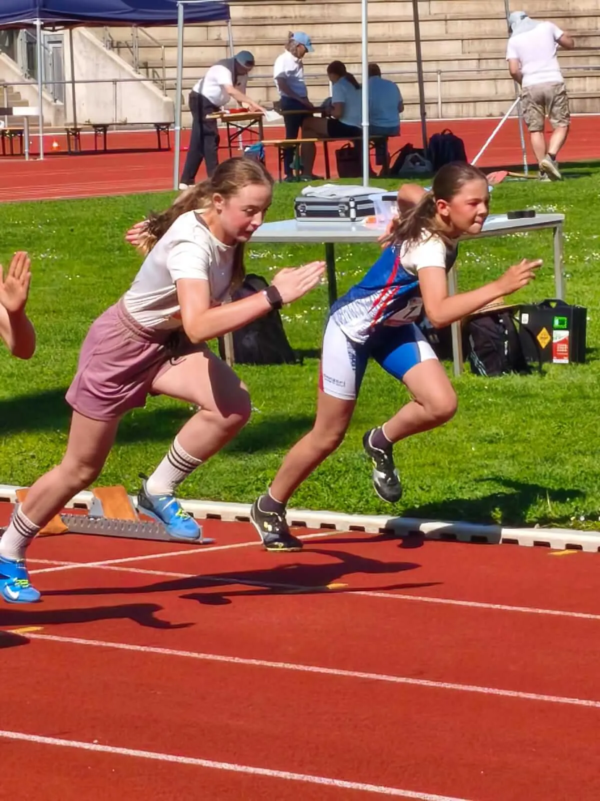 Zwei junge Athletinnen in Startposition auf einer roten Laufbahn, bereit für einen Sprint. Im Hintergrund sind weitere Sportler und Zuschauer zu sehen.