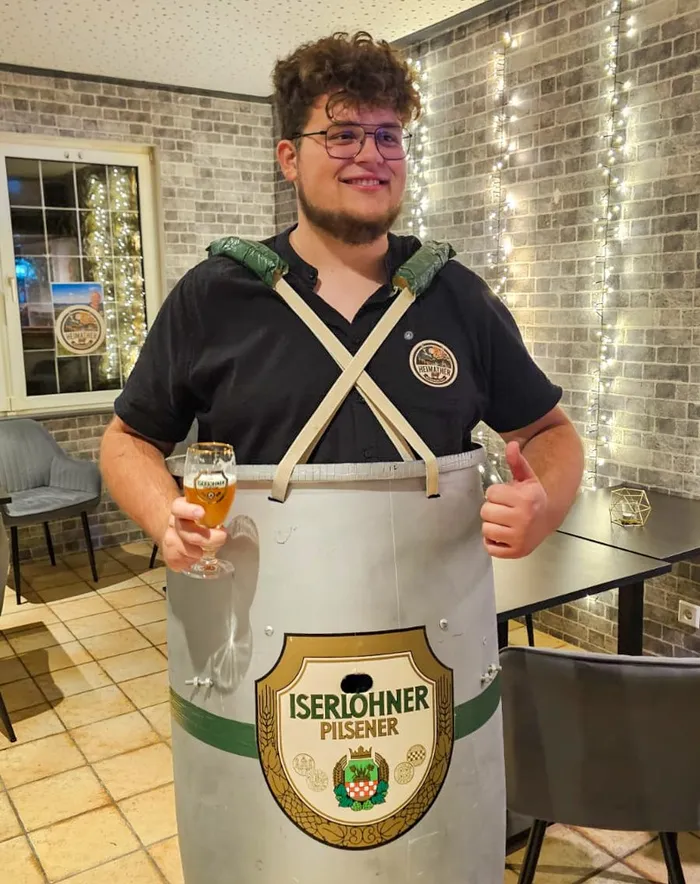 Ein junger Mann in einem Kostüm aus einer silbernen Iserlohner Pilsener Tonne hält ein Glas Bier und zeigt einen Daumen nach oben.