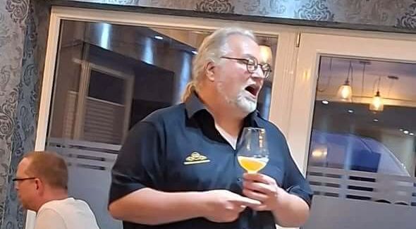 Biersommelier Markus Raupach hält ein Glas Bier und spricht mit offenem Mund in einem Raum mit dekorativen Wänden.