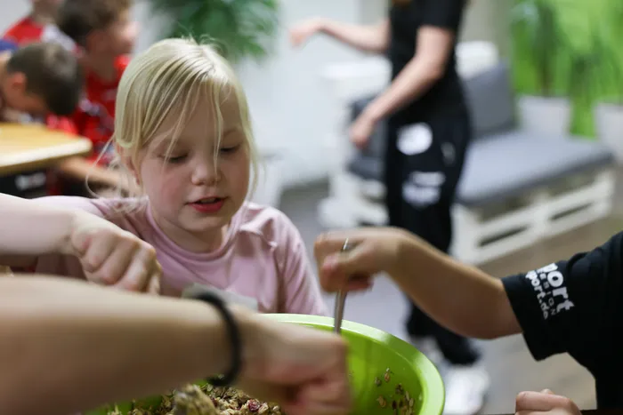 Ein Mädchen mit blonden Haaren rührt mit einem Löffel in einer Schüssel mit Müsli. Eine Hand reicht ihr Zutaten zu. Im Hintergrund sind weitere Kinder und Erwachsene zu sehen.