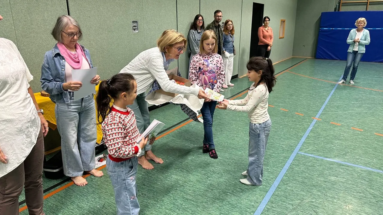 Eine Frau überreicht einem Mädchen in einer Turnhalle ein Buch, während andere Kinder und Erwachsene zusehen.