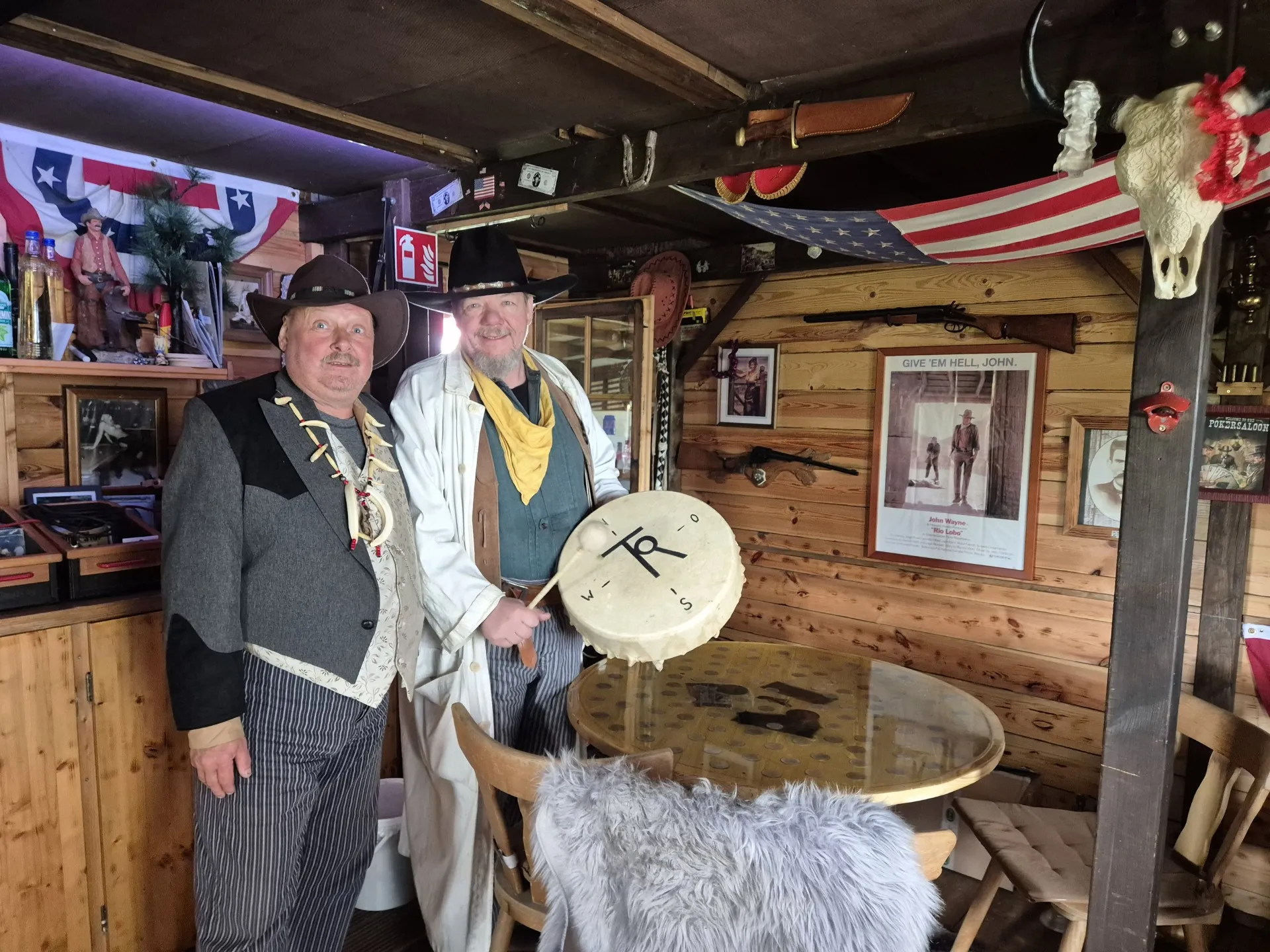 Zwei Männer mit Cowboyhüten und Westernkleidung stehen lächelnd vor einer Bar mit Western-Dekoration und amerikanischer Flagge.