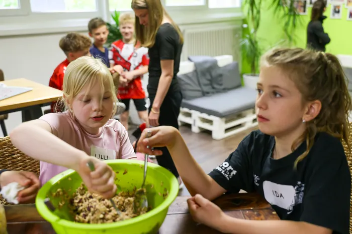 Zwei Mädchen, Lilli und Ida, rühren in Schüsseln mit einer Mischung aus Zutaten. Im Hintergrund bereiten weitere Kinder mit einer Betreuerin gesunde Snacks zu.