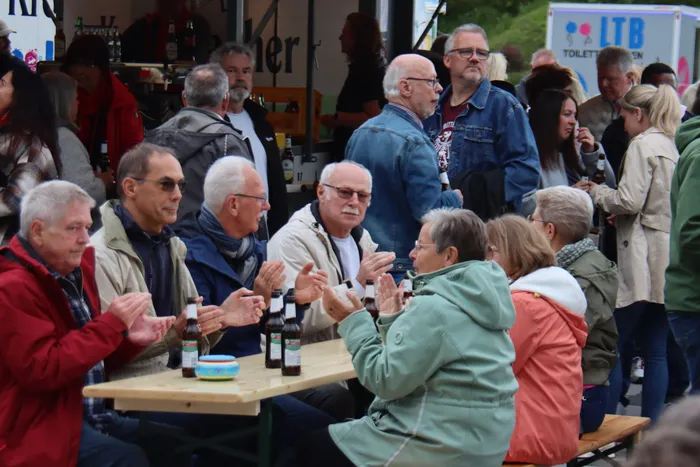 Eine Gruppe von Menschen sitzt an einem Tisch im Freien und klatscht, während im Hintergrund weitere Menschen stehen und sich unterhalten. Bierflaschen stehen auf dem Tisch.