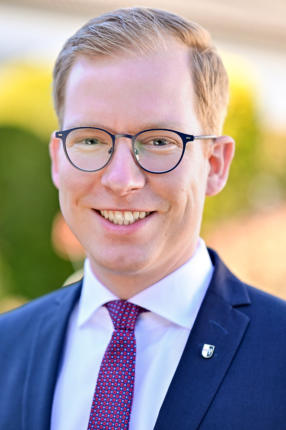 Christian Schweitzer lächelt in einem Porträt mit Brille, weißem Hemd und roter Krawatte.