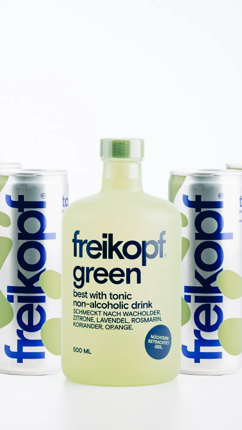 Freikopf Green alkoholfreies Getränk in einer Flasche vor weiteren Flaschen des gleichen Produkts. Auf dem Etikett stehen die Geschmacksrichtungen Wacholder, Zitrone, Lavendel, Rosmarin, Koriander und Orange.