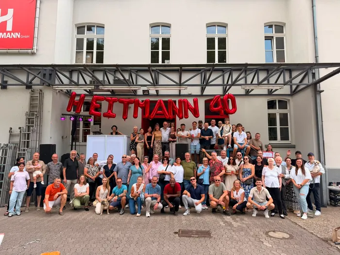 Eine große Gruppe von Menschen posiert vor einem weißen Gebäude unter großen roten aufblasbaren Buchstaben mit der Aufschrift „HEITMANN 40“.