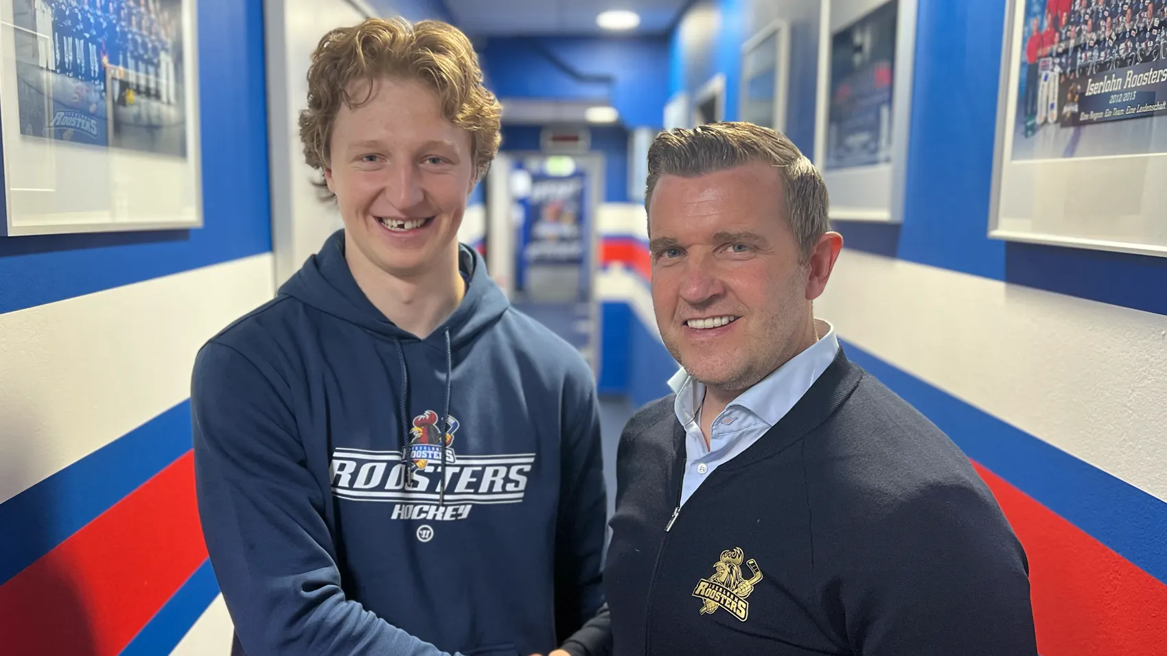 Zwei Männer, Roosters-Sportdirektor Franz-David Fritzmeier und Neuzugang Norwin Panocha, stehen lächelnd in einem Gang der Eissporthalle am Seilersee.
