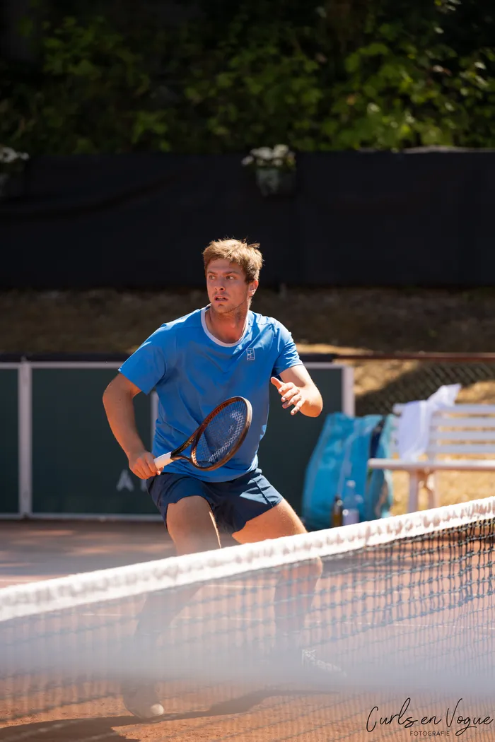 Ein Tennisspieler in blauem T-Shirt und dunklen Shorts steht mit einem Schläger auf einem Sandplatz vor dem Netz.