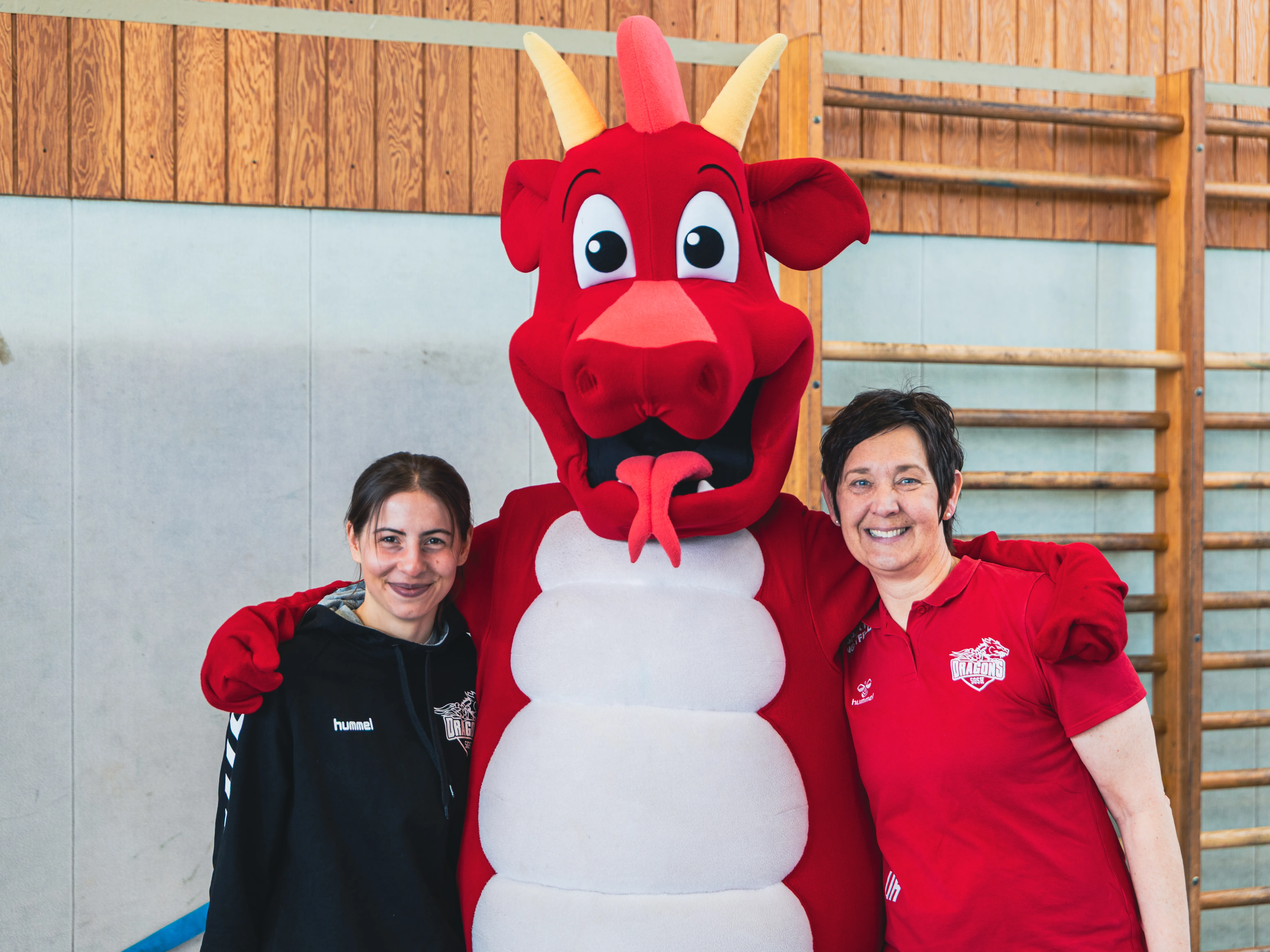Zwei Frauen umarmen ein rot gekleidetes Drachenmaskottchen in einer Turnhalle. Im Hintergrund sind Holzbalken und eine Wand mit Sprossen zu sehen.
