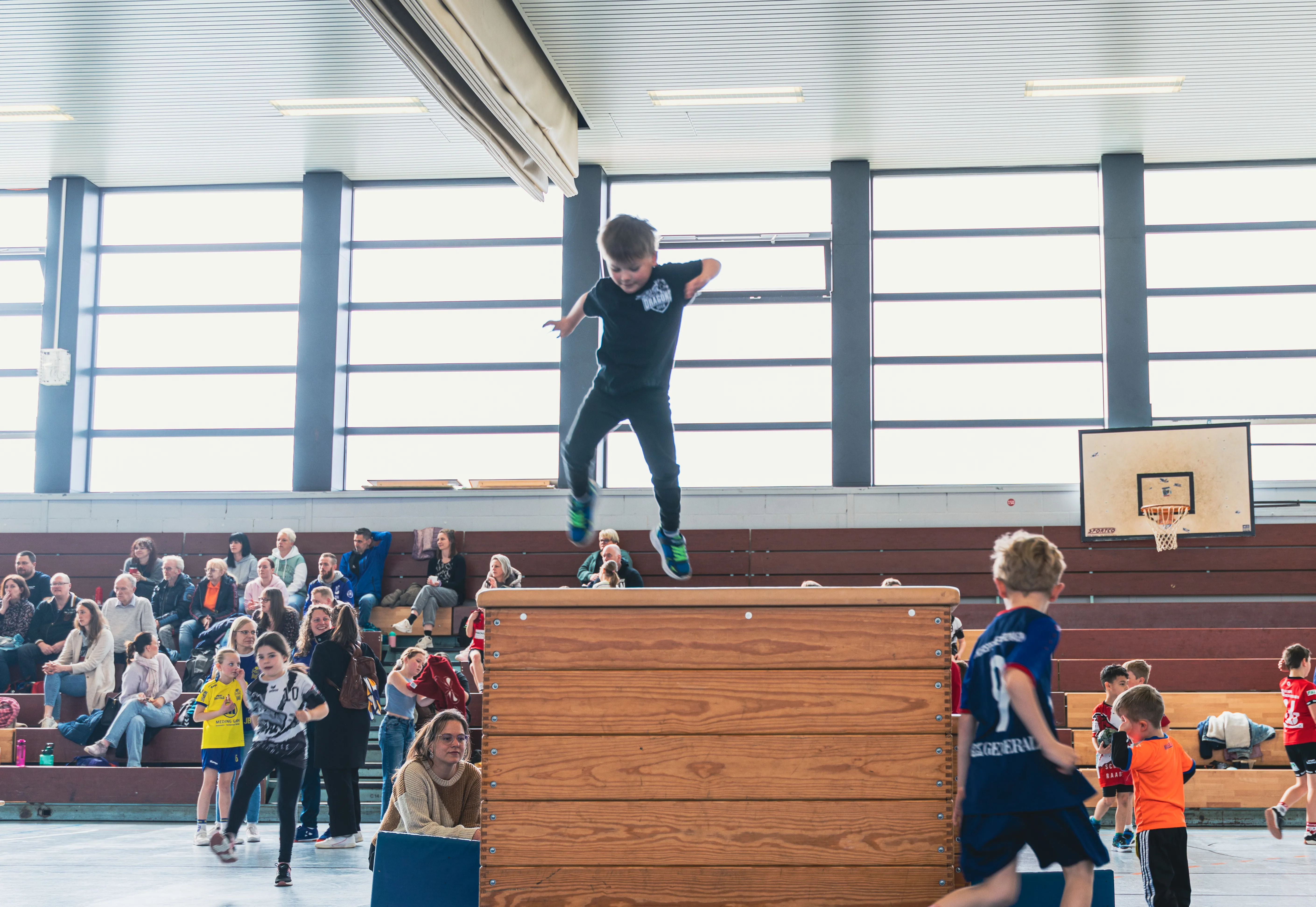 Ein Junge springt über eine hölzerne Kiste, während andere Kinder und Zuschauer im Hintergrund zusehen.