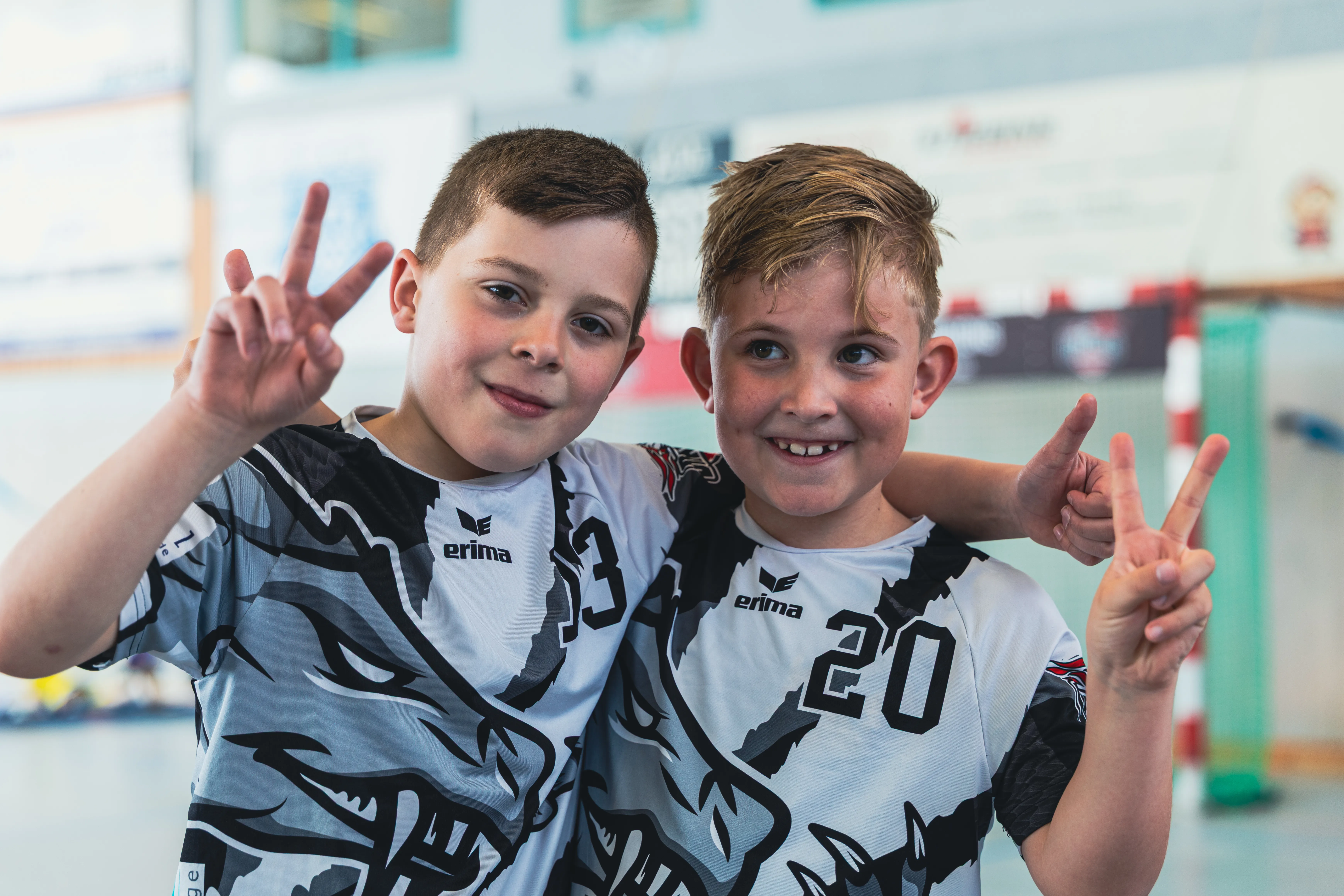 Zwei lächelnde Jungen der F-Jugend der SGSH Dragons zeigen den Friedenszeichen vor einer Sporthalle.