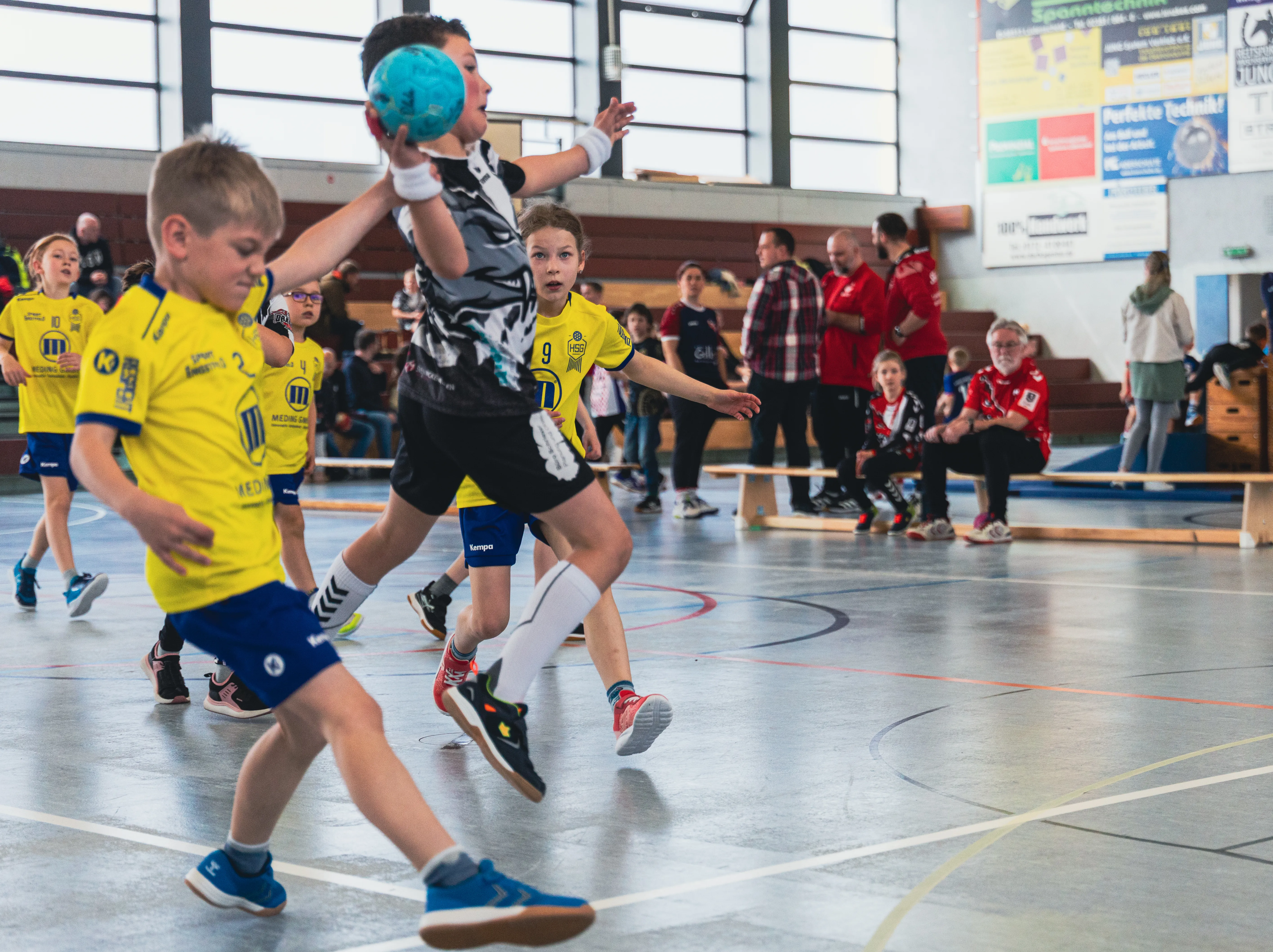 Handballspieler der F-Jugend im aktiven Spiel während eines Spielfestes, wobei ein Spieler den Ball wirft und ein anderer ihn fängt. Im Hintergrund stehen Zuschauer in einer Sporthalle.