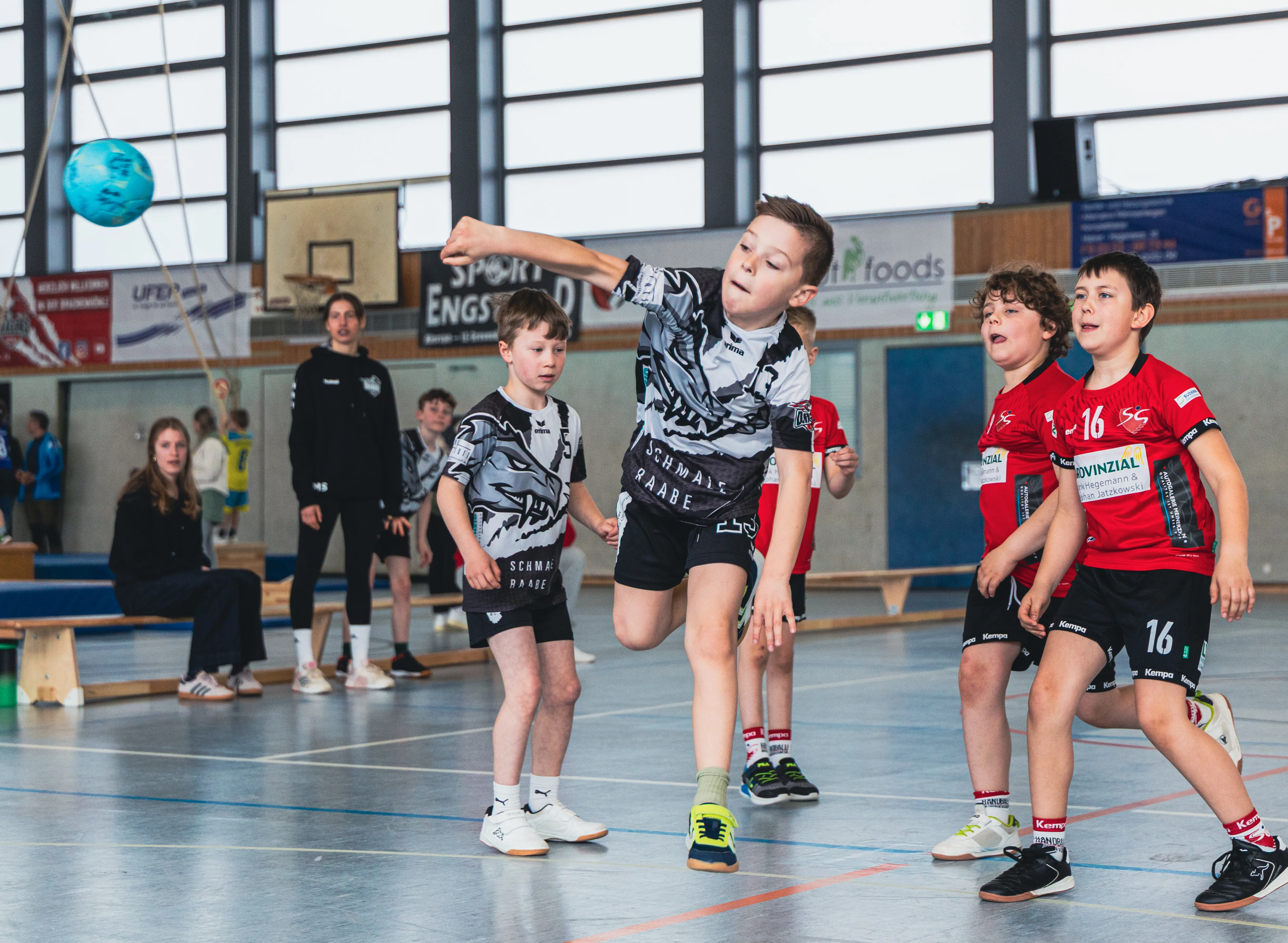 Junge Handballspieler werfen und fangen den Ball während eines Spiels in einer Sporthalle. Im Hintergrund stehen weitere Kinder und Erwachsene.