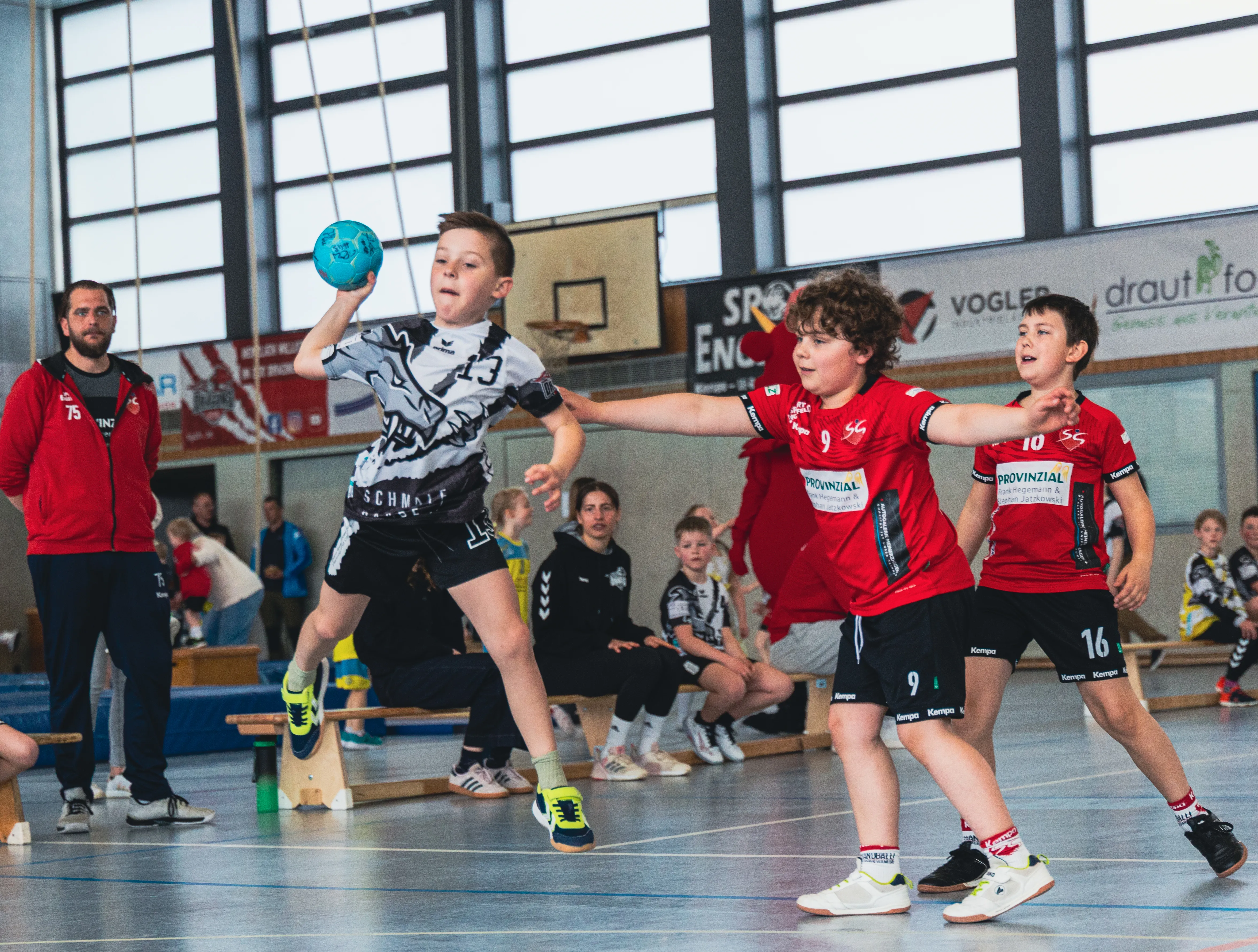 Ein Jugendhandballspieler wirft den Ball während eines Spiels. Im Hintergrund stehen weitere Spieler und Zuschauer in einer Sporthalle.