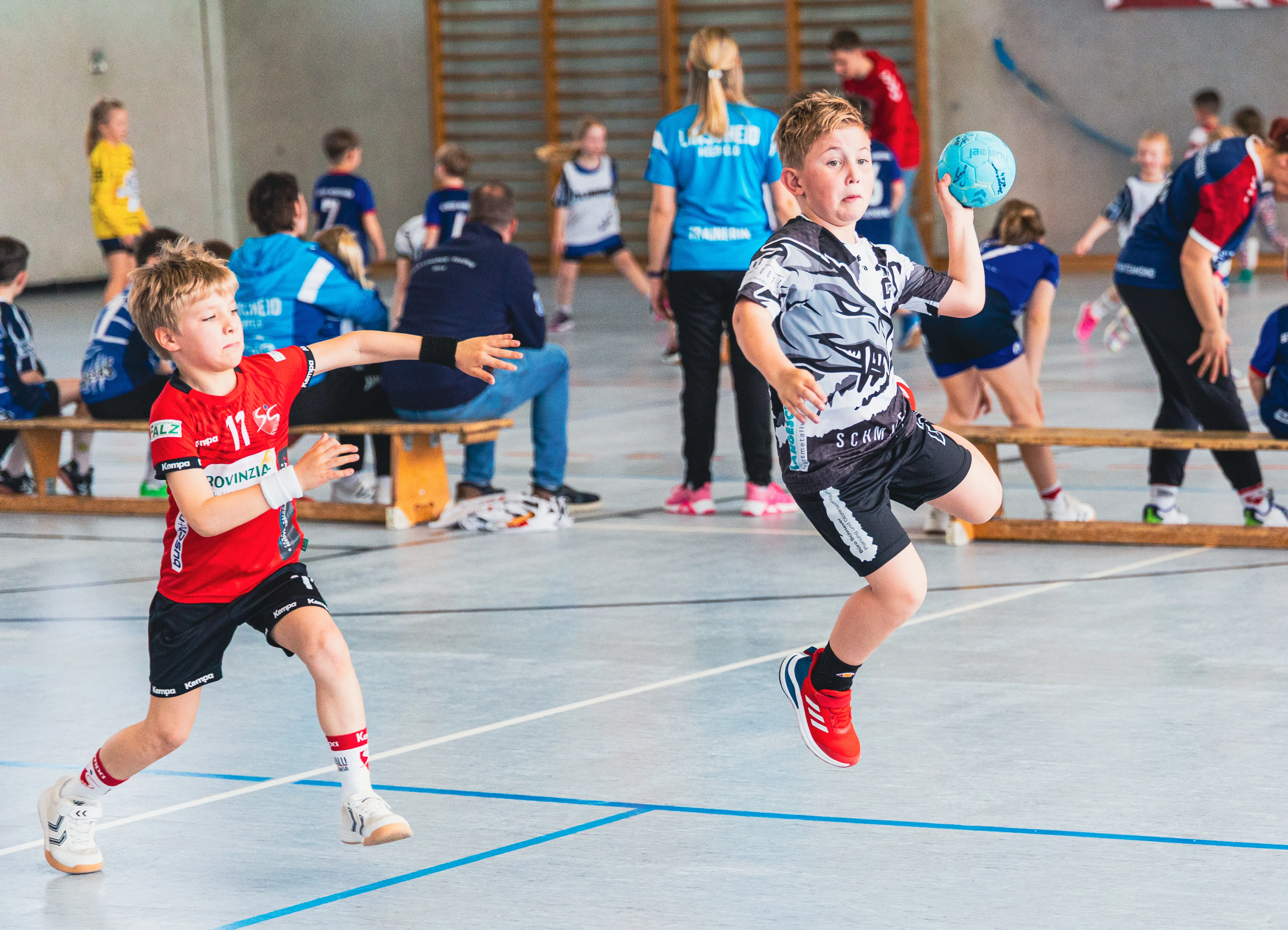 Zwei junge Handballspieler im Spiel, einer in rotem Trikot springt zum Wurf, der andere in grau-weißem Trikot versucht den Ball zu blocken.