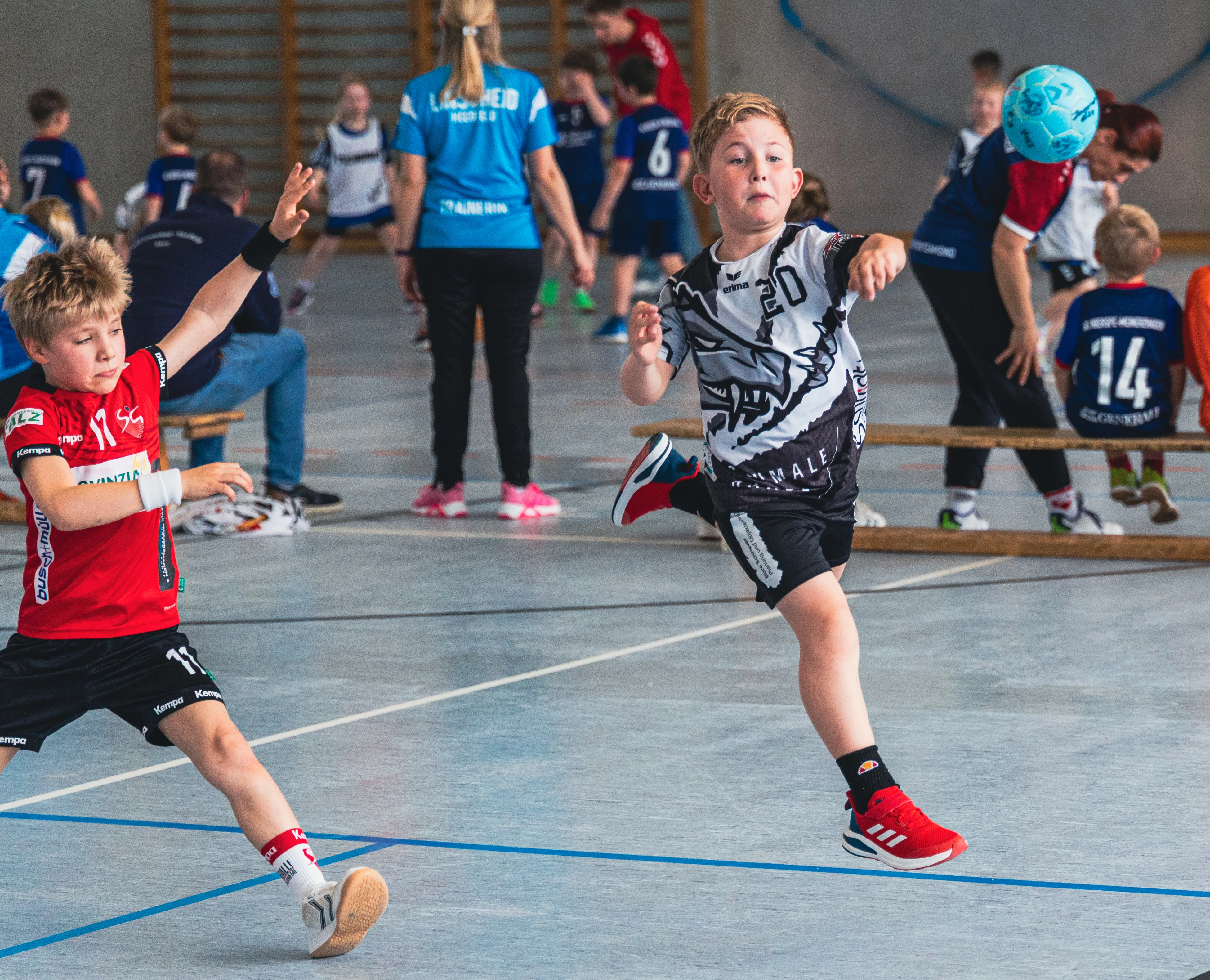 Zwei Jungen spielen Handball in einer Sporthalle. Der Junge links trägt ein rotes Trikot, der andere ein schwarz-weißes.