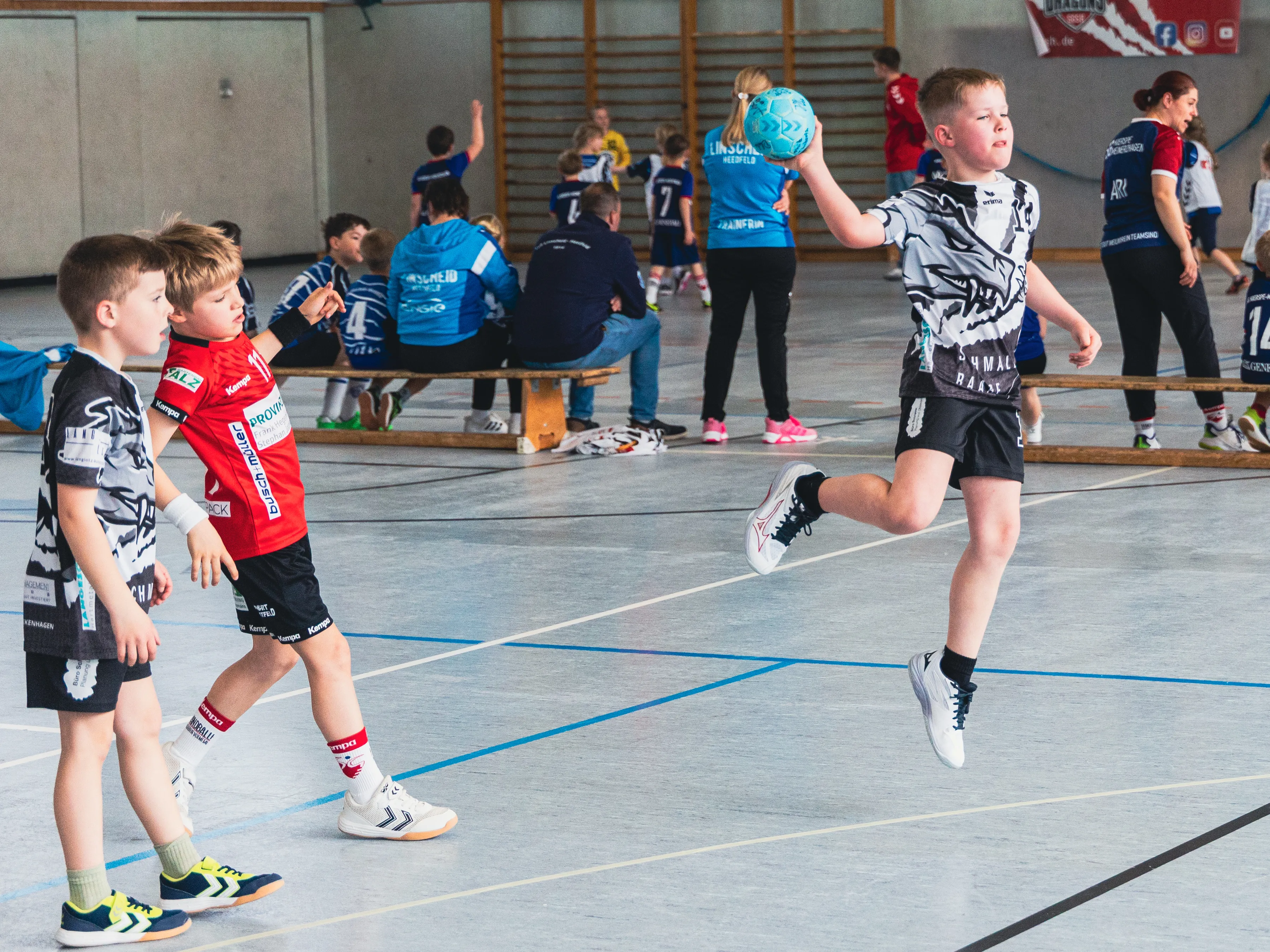 Zwei Jungen spielen Handball in einer Sporthalle, während im Hintergrund weitere Kinder trainieren.