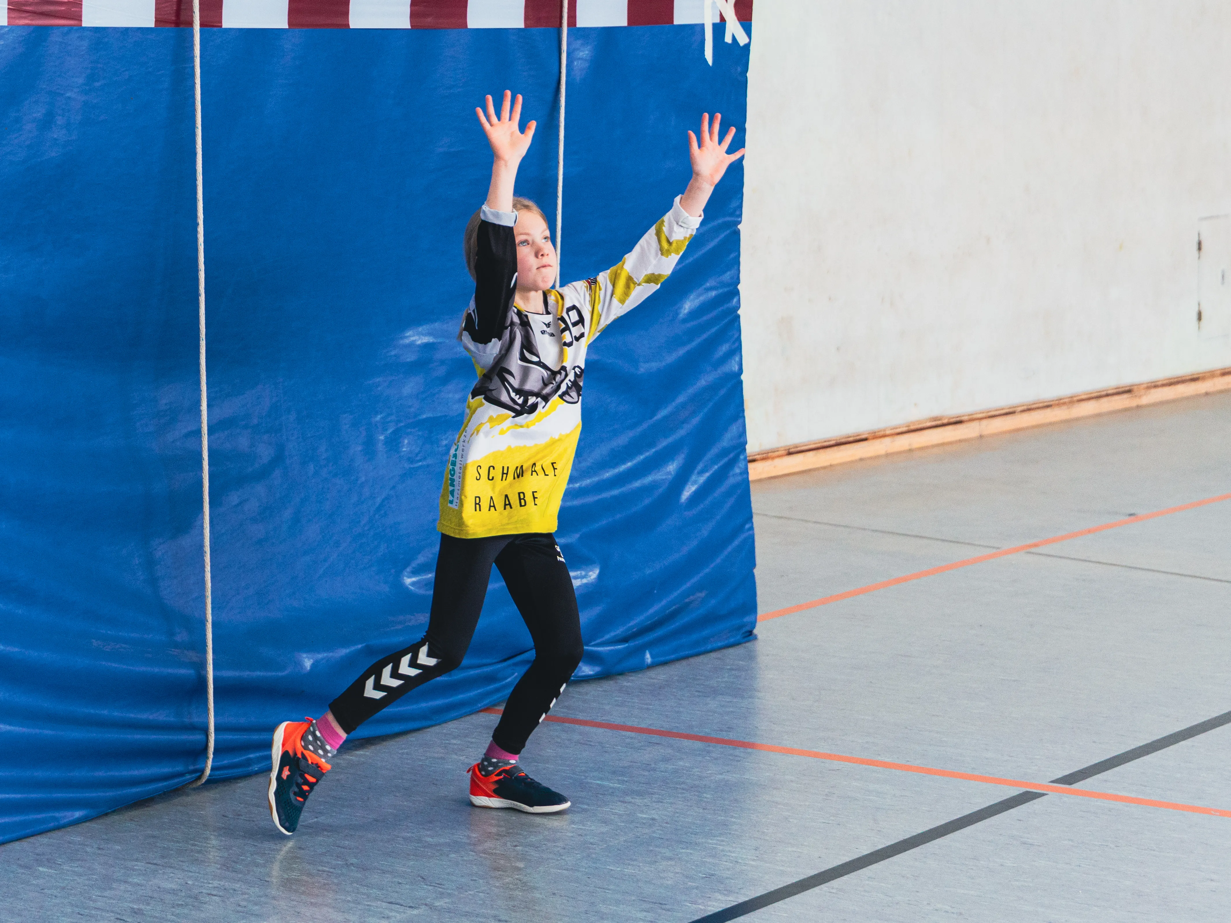 Junge Handballspielerin springt mit erhobenen Armen vor blauem Torwartwand in Sporthalle.