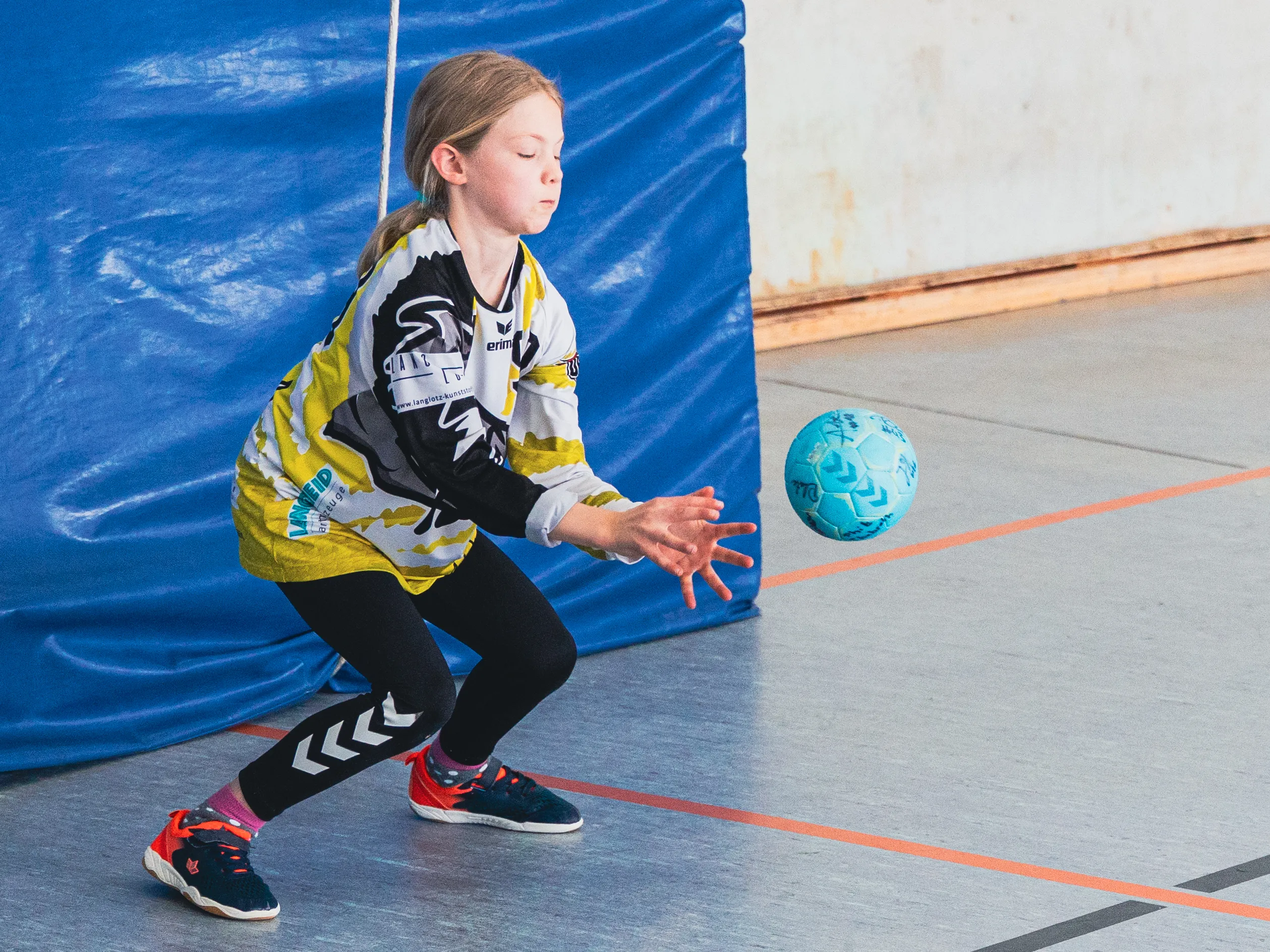 Ein Mädchen in Handballkleidung wirft den Ball während eines Spiels. Im Hintergrund eine blaue Wand mit Markierungen.