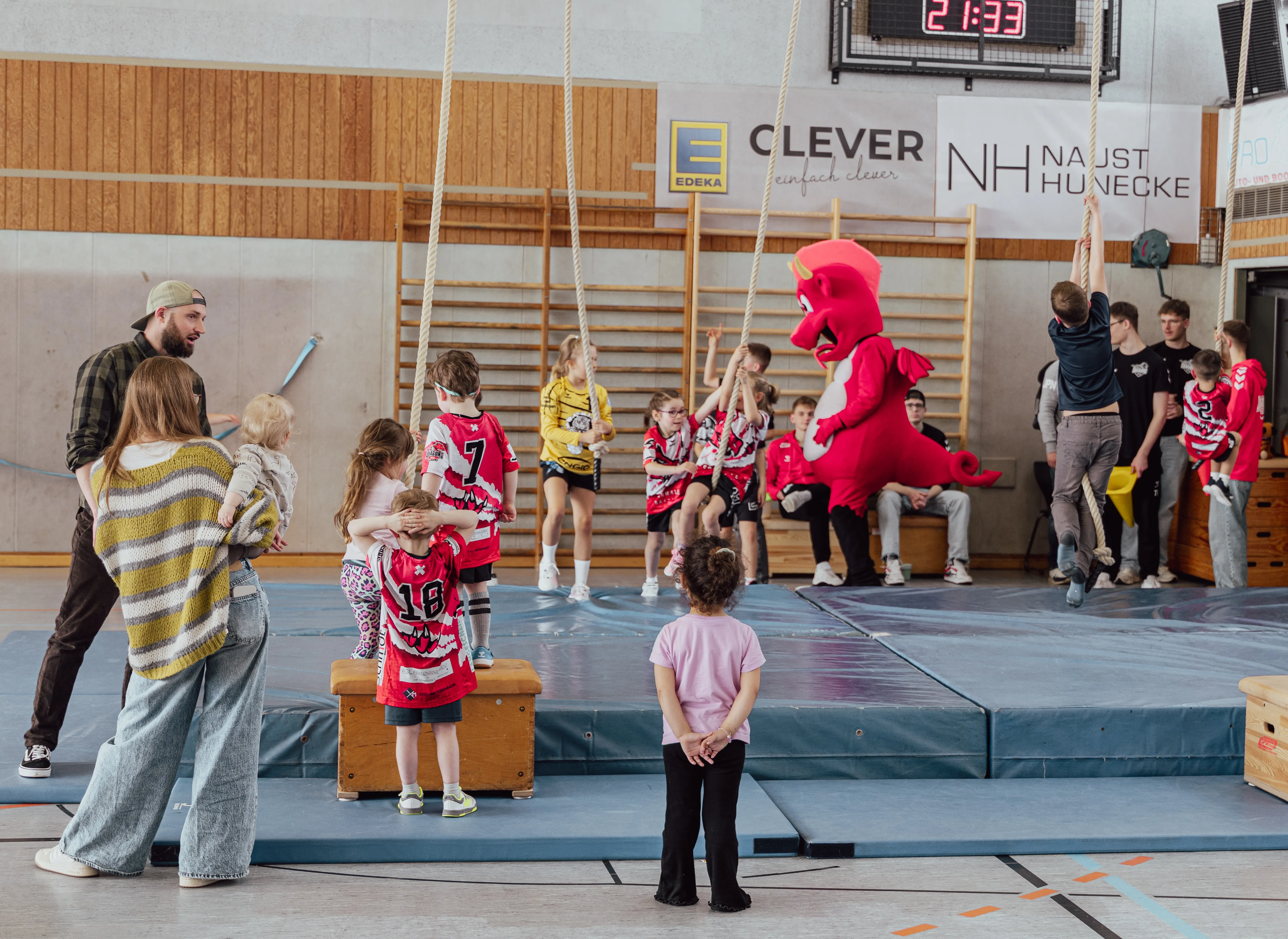 Mehrere Kinder in roten Trikots stehen und sitzen in einer Turnhalle um eine Schaukel. Ein Mann mit Bart und eine Frau beobachten die Kinder, während ein rotes Drachenmaskottchen neben den Kindern steht.