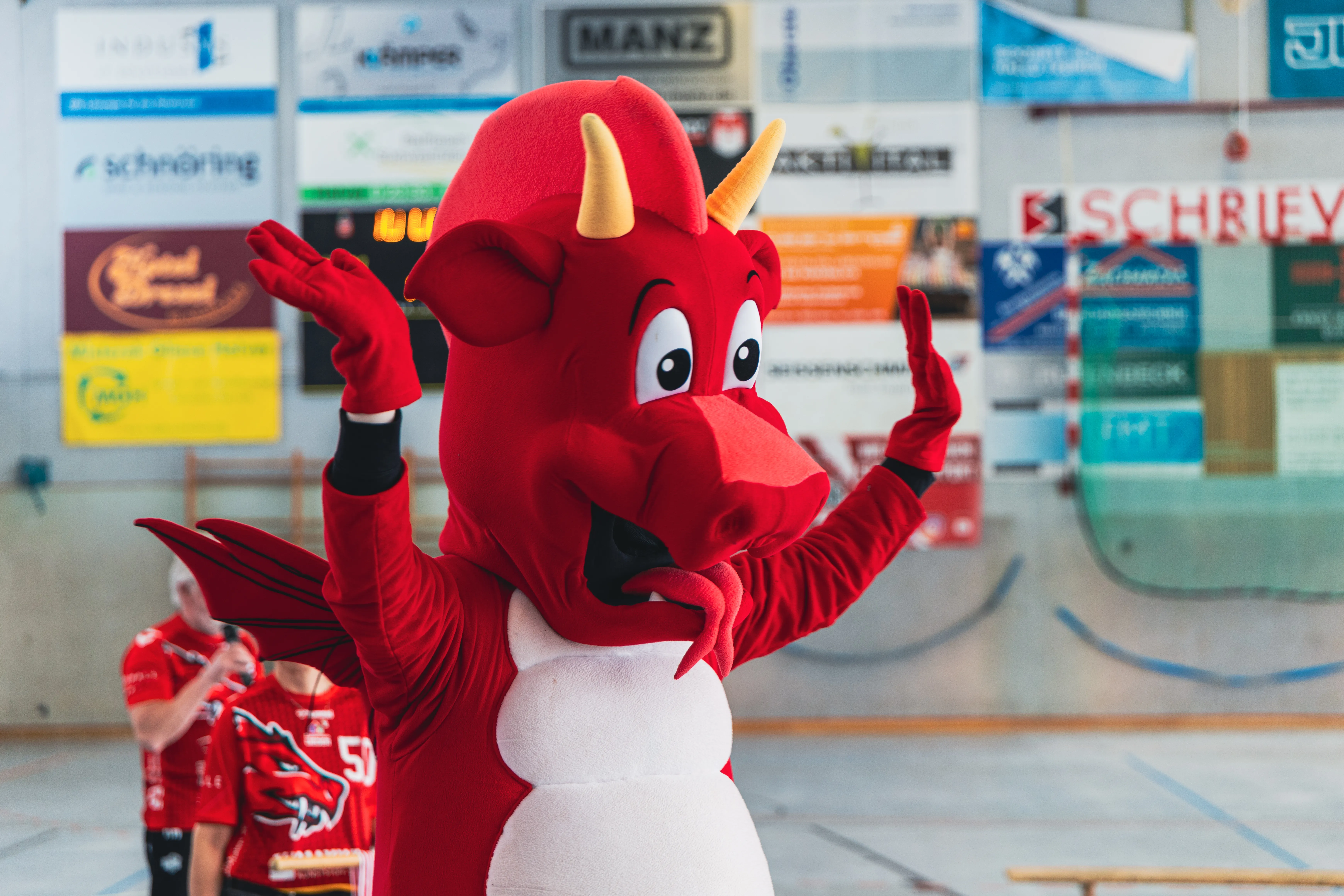 Rotes Drachenmaskottchen mit gelben Hörnern und großen Augen steht mit erhobenen Armen in einer Sporthalle. Im Hintergrund sind Werbebanner und verschwommen spielende Kinder zu sehen.