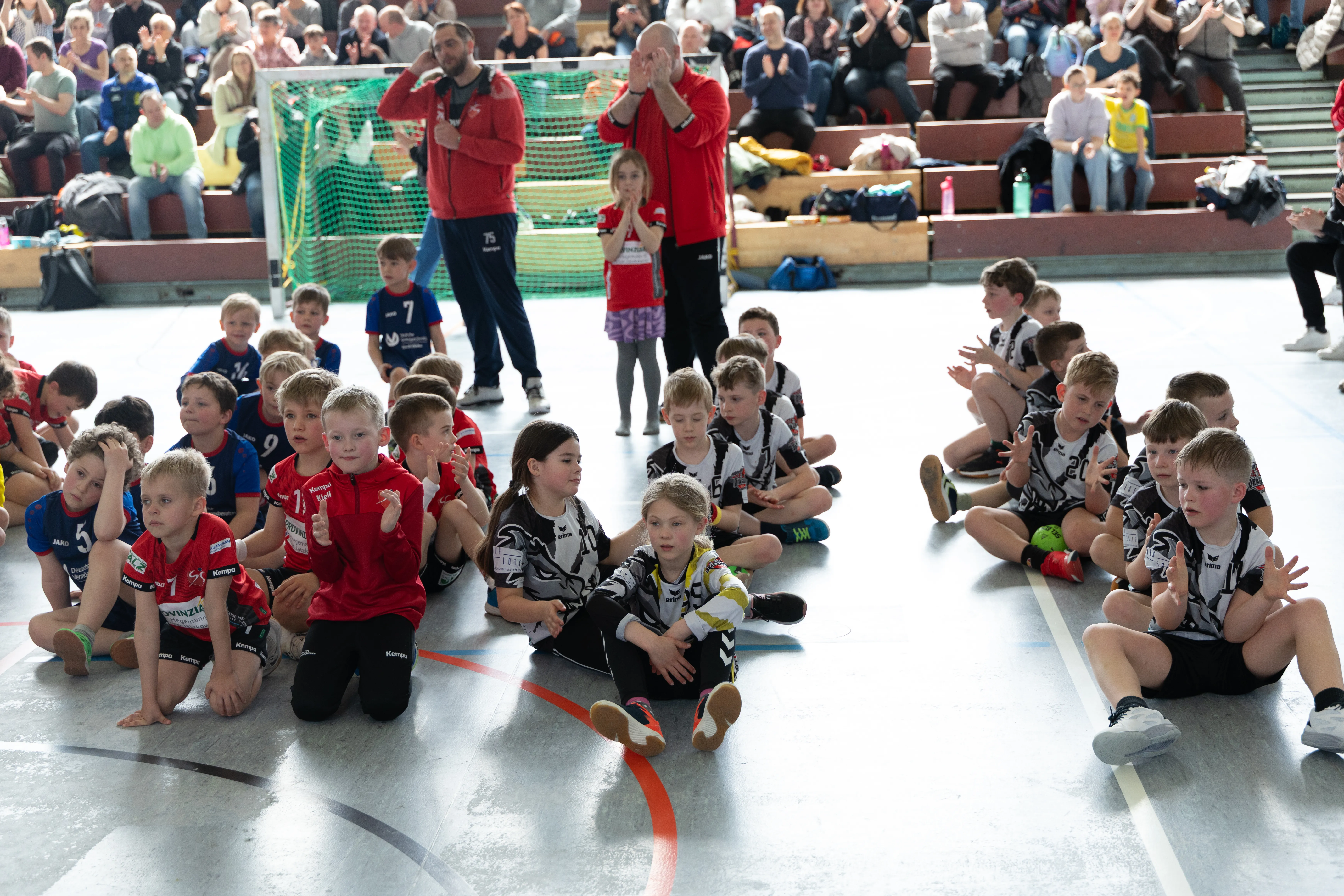 Eine Gruppe von Kindern im Handballtrikot sitzt im Kreis auf einer Sporthalle und schaut erwartungsvoll nach vorne. Im Hintergrund stehen und sitzen weitere Kinder und Erwachsene und beobachten die Szene.