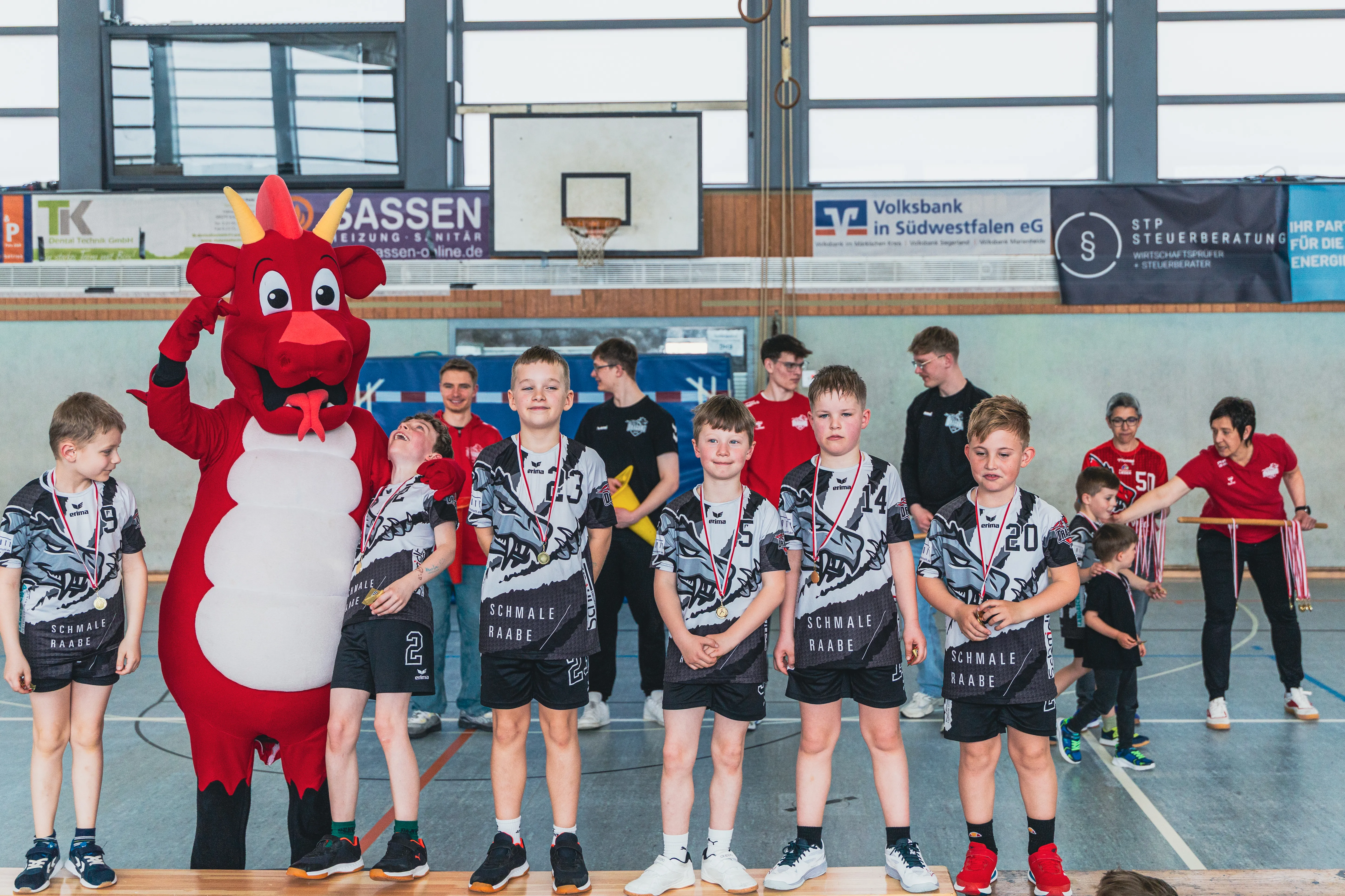 Eine Gruppe Jugendlicher in Handballtrikots mit dem Schriftzug „Schmale Raabe“ steht mit Medaillen um den Hals in einer Sporthalle. Neben ihnen steht ein rotes Drachenmaskottchen und hebt die Hand.