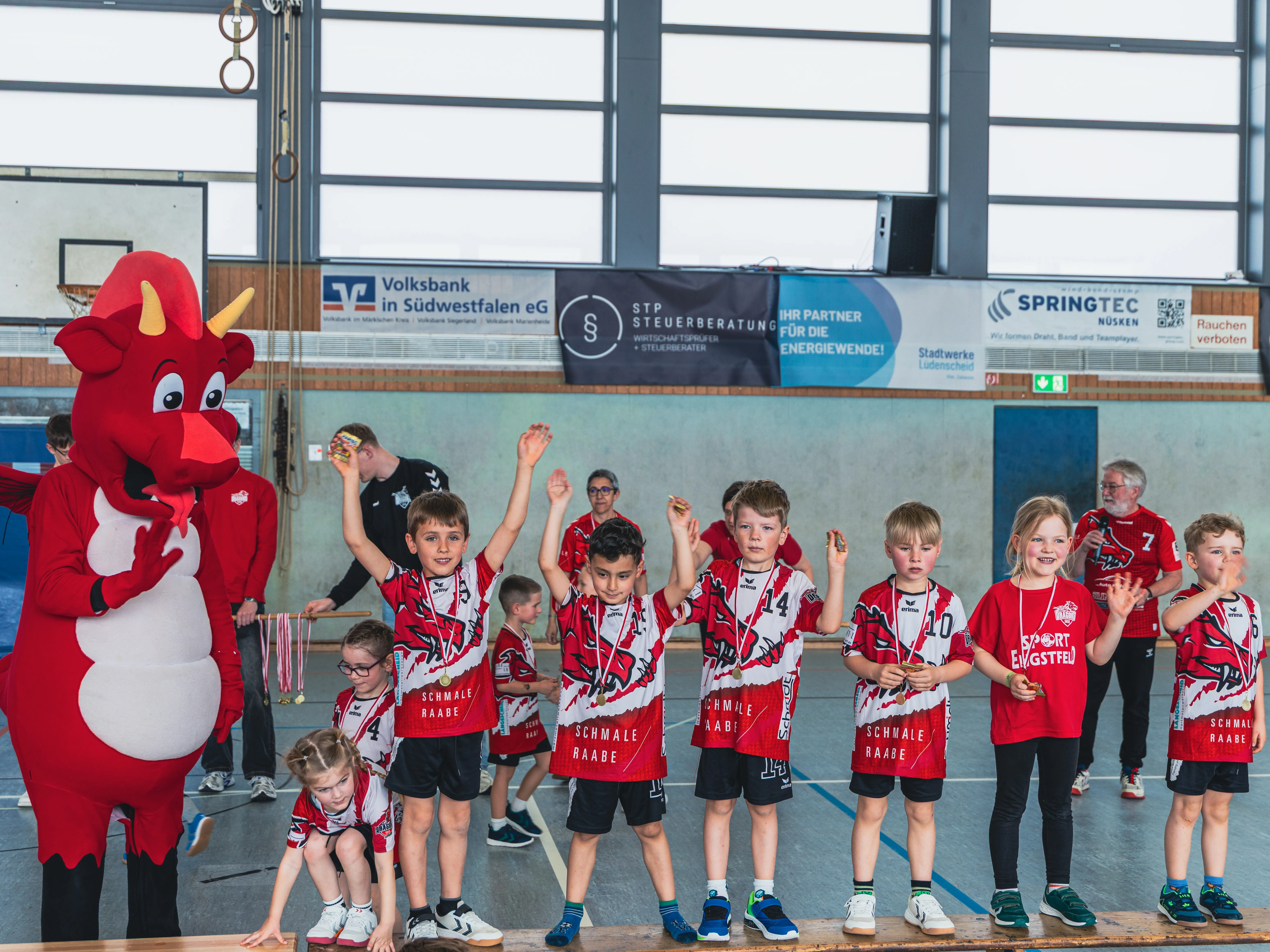 Eine Gruppe von Kindern in Handballtrikots der SGSH Dragons steht mit Medaillen um den Hals und jubelt. Neben ihnen steht ein rotes Maskottchen in Form eines Drachen.