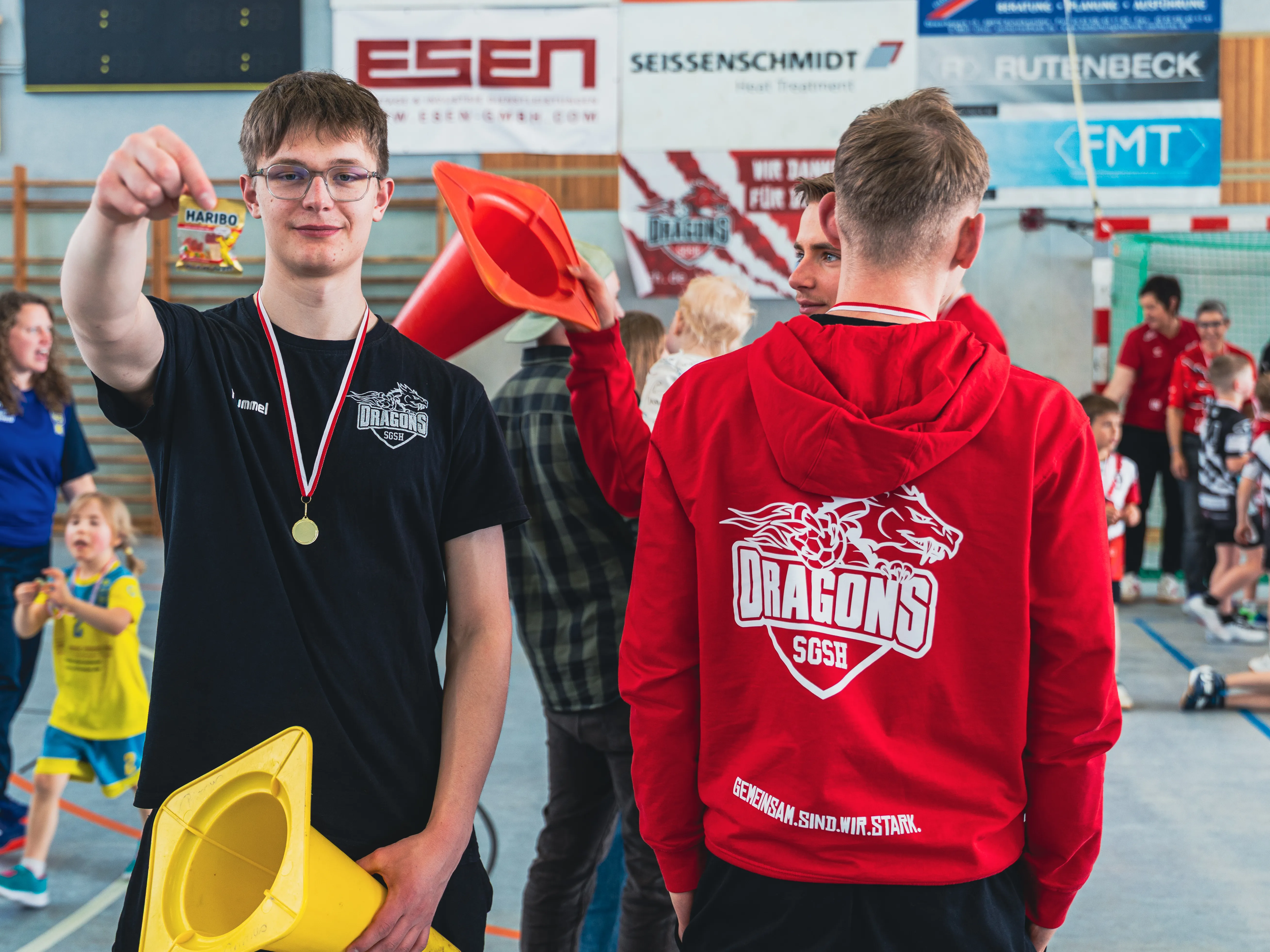 Zwei junge Handballspieler stehen in einer Sporthalle, einer trägt ein rotes Jackett mit dem Aufdruck „Dragons“, der andere ein schwarzes T-Shirt. Im Hintergrund sind weitere Kinder und Zuschauer zu sehen.