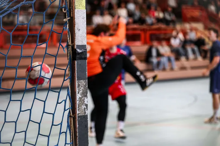 Ein rot-weißer Handball im blauen Tornetz vor einem springenden Torwart in orangefarbenem Trikot.
