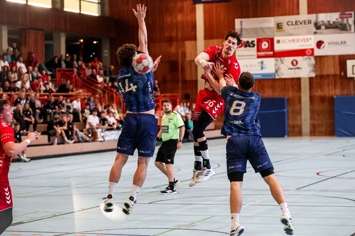 Ein Handballspieler in einem roten Trikot springt in der Luft, während er von zwei Spielern in blauen Trikots verteidigt wird.