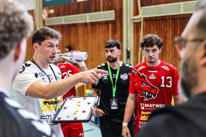 Trainer Jens Gawer gibt mit einem Taktikbrett taktische Anweisungen an seine Spieler in roten Trikots.