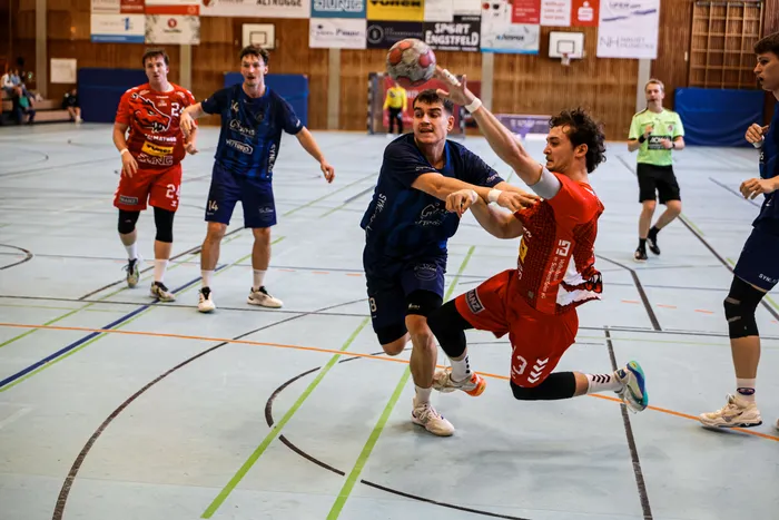 Ein Handballspieler in einem blauen Trikot wird von einem Spieler in einem roten Trikot beim Sprungwurf blockiert.