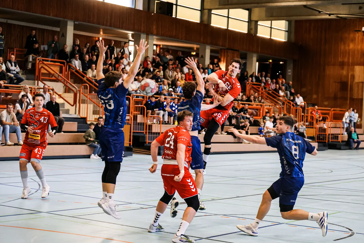 Ein Handballspieler in einem roten Trikot springt zu einem Wurf, während er von Spielern in blauen Trikots in einer Sporthalle verteidigt wird.