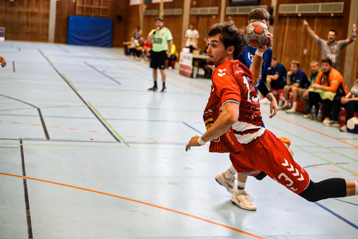 Ein Handballspieler in einem roten Trikot springt mit dem Ball in der Hand in einer Sporthalle.