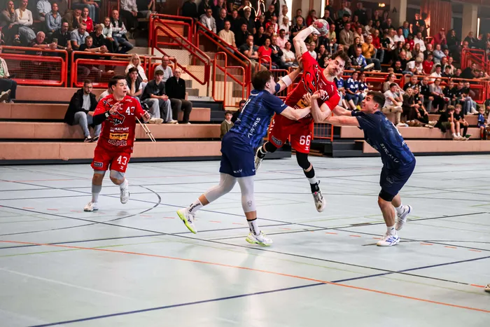 Ein Handballspieler in rotem Trikot springt zum Wurf, während zwei Gegenspieler in blauen Trikots ihn am Sprung abfangen.