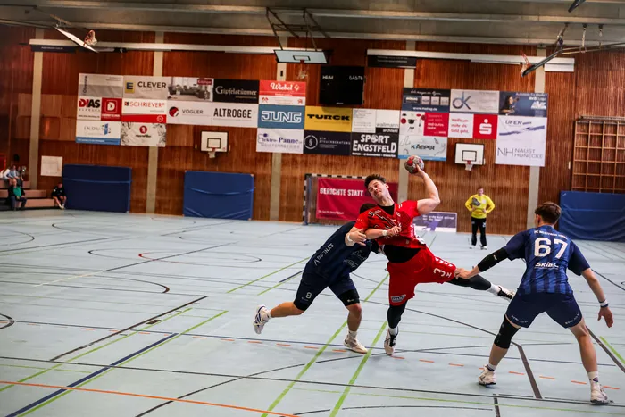 Ein Handballspieler in einem roten Trikot springt mit dem Ball in der Hand, während er von einem Spieler in einem dunkelblauen Trikot bedrängt wird.