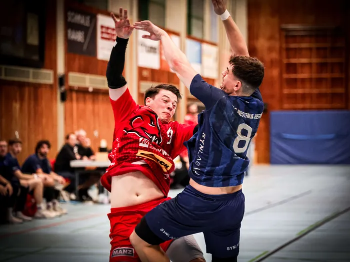 Ein Handballspieler in einem roten Trikot und ein Spieler in einem blauen Trikot mit der Nummer 8 springen in einer Sporthalle hoch.