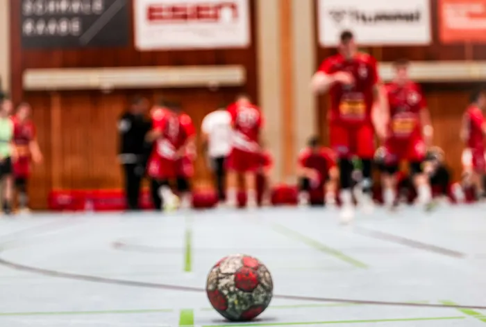 Ein Handball liegt im Vordergrund auf dem Hallenboden, im Hintergrund sind verschwommen Spieler in roten Trikots zu sehen.