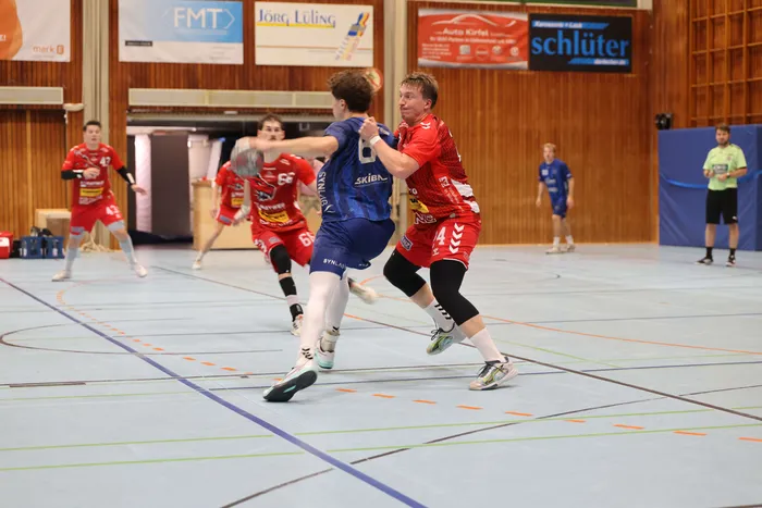 Ein Handballspieler in einem blauen Trikot mit dem Ball wird von einem Spieler in einem roten Trikot während eines Spiels in einer Sporthalle verteidigt.