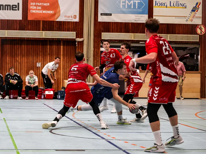 Handballspieler in roten und blauen Trikots im Zweikampf während eines Spiels in einer Sporthalle.