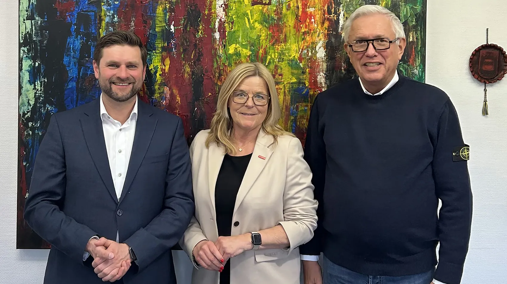 Hendrik Schmitt, Uta Neumeister und Bürgermeister Ralf Beßler stehen vor einem abstrakten Gemälde zusammen. Schmitt und Beßler tragen Anzug, Neumeister eine hellgraue Jacke.