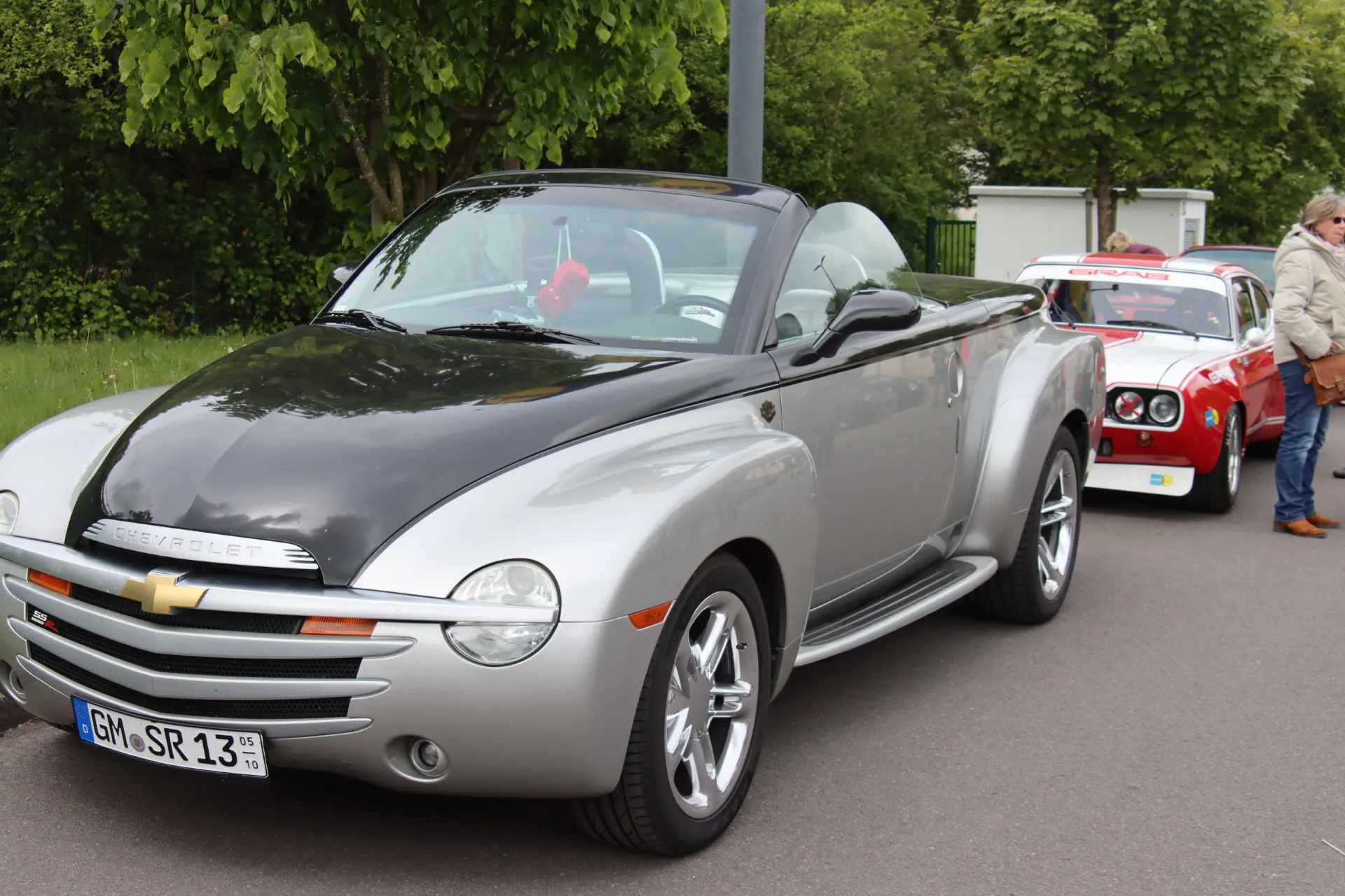 Silberner Chevrolet SSR mit offener Karosserie und schwarzem Motorhaubenbereich steht im Fokus, während im Hintergrund ein rotes Oldtimer-Auto und Personen zu sehen sind.