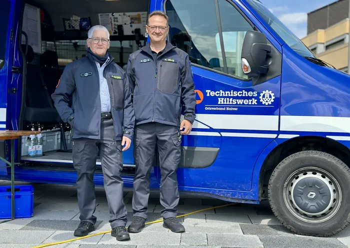 Zwei Männer in dunkelblauen Uniformen des Technischen Hilfswerks stehen vor einem blauen THW-Transporter.